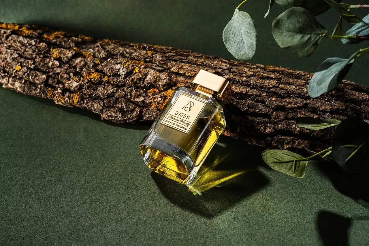 Eleganckie perfumy B. Bates Oriental Woody, które zostawiają ogon, leżą obok kawałka kory drzewa, otoczone zielonymi liśćmi.
