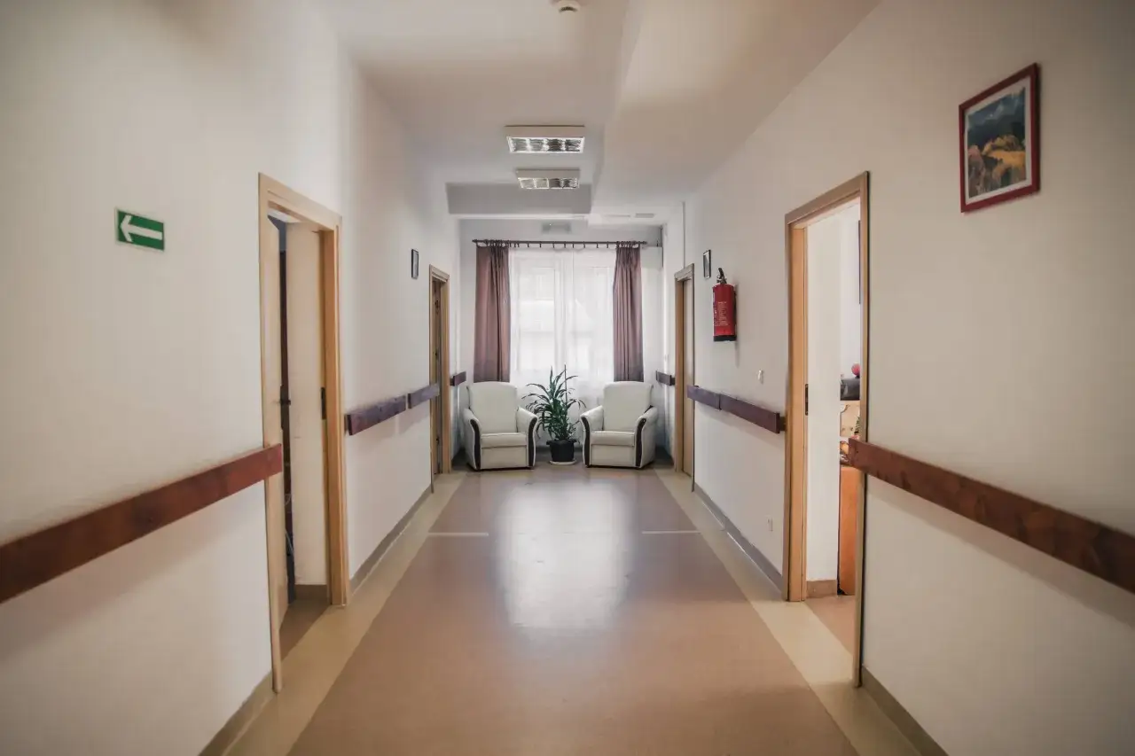 Jasny korytarz zakładu opiekuńczo-leczniczego z poręczami, fotelami i roślinami. Sprawdź wolne miejsca.