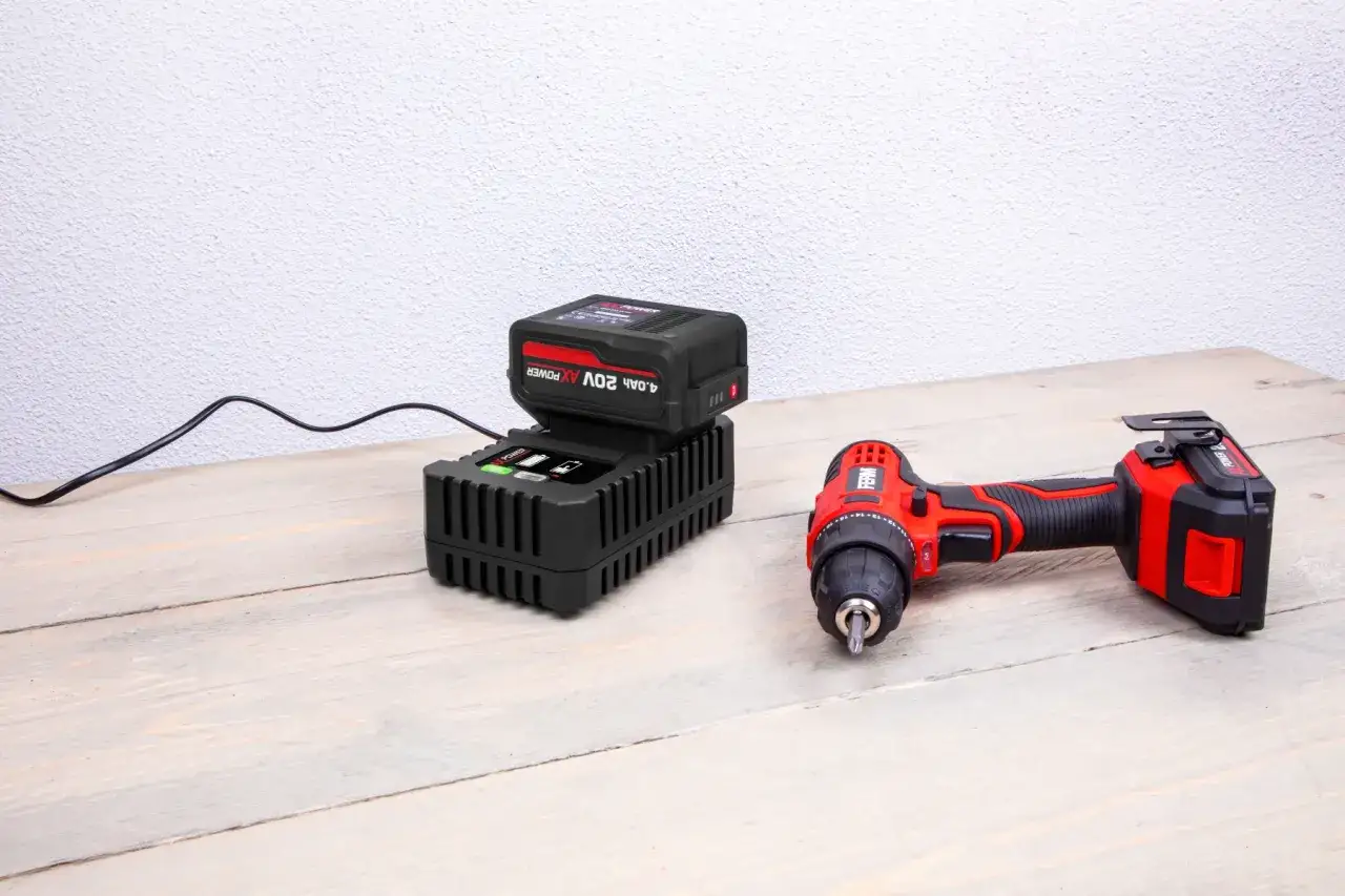 Une batterie 4Ah est en charge sur un chargeur 2Ah. Une perceuse sans fil rouge et noire est à côté.