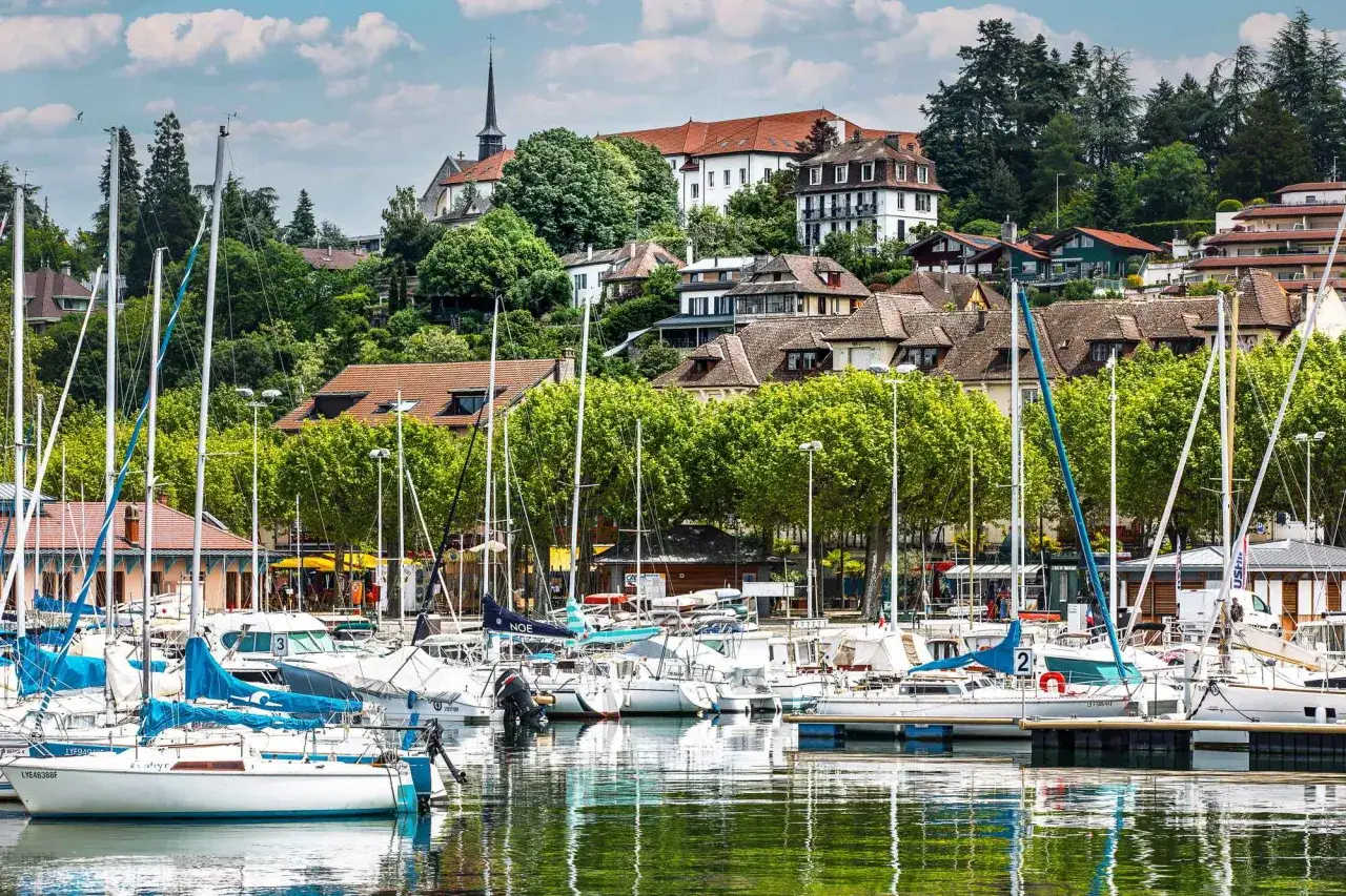 Port de plaisance à Thonon-les-Bains, avec des voiliers blancs et bleus amarrés, des maisons sur la colline et une église. Idéal pour savoir que faire à Thonon-les-Bains.