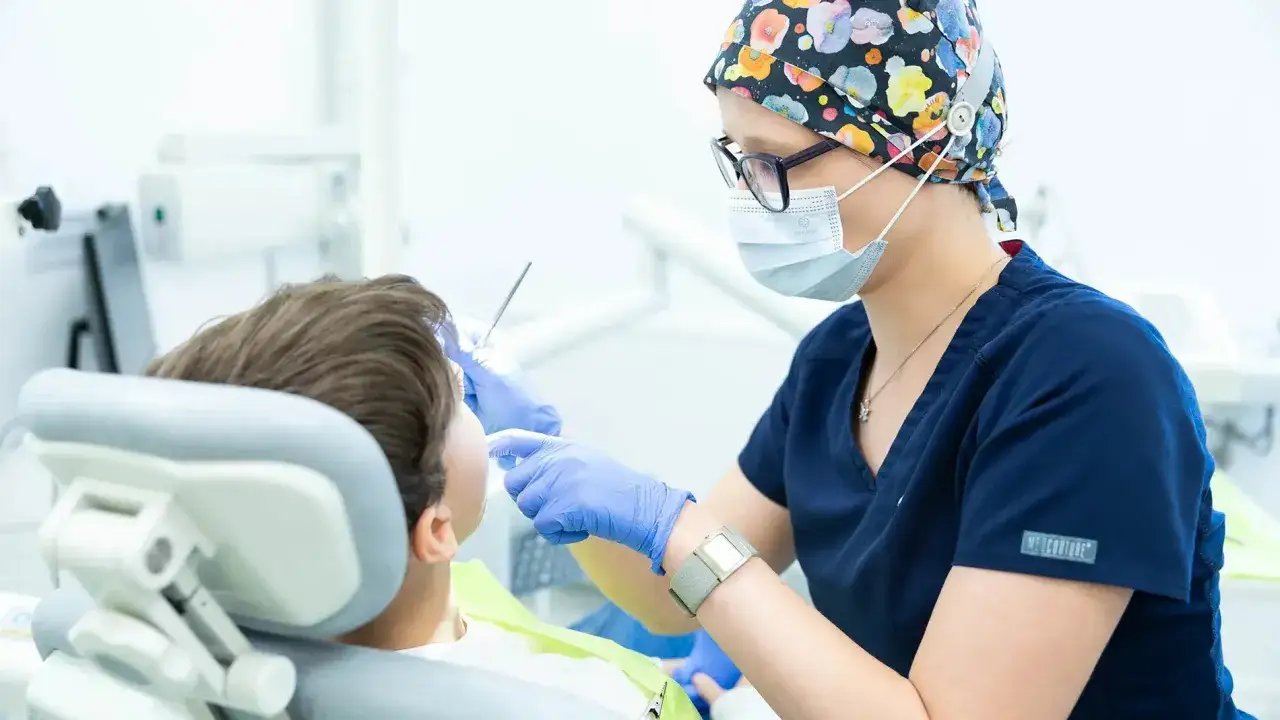 Dentystka w masce i kolorowym czepku bada zęby pacjenta siedzącego na fotelu dentystycznym.