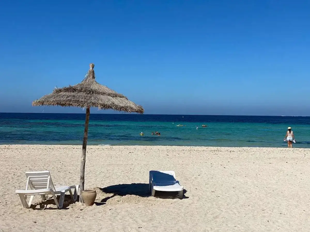 Słoneczne tunezyjskie plaże, turkusowe morze, leżaki pod strzechą i kobieta w białej sukience. Idealne wakacje!