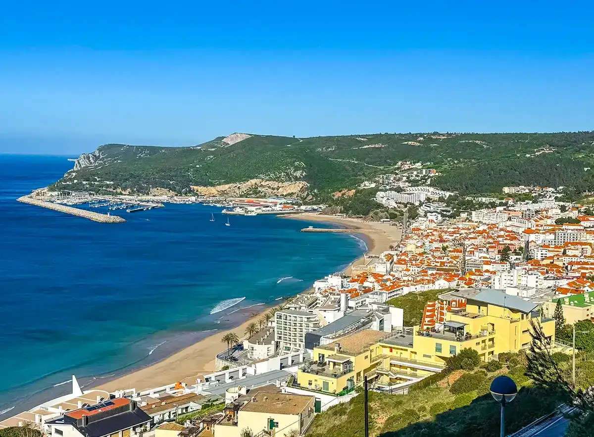 Blick auf die Küste von Sesimbra, Portugal, mit einem malerischen Strand, einem geschäftigen Hafen und weiß getünchten Häusern mit roten Ziegeldächern.