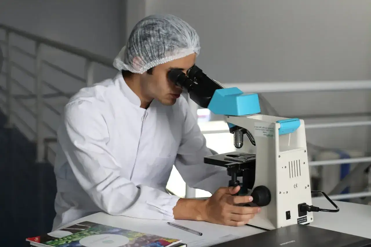 Młody technik farmacji w białej kurtce i czepku pochyla się nad mikroskopem, badając próbki.
