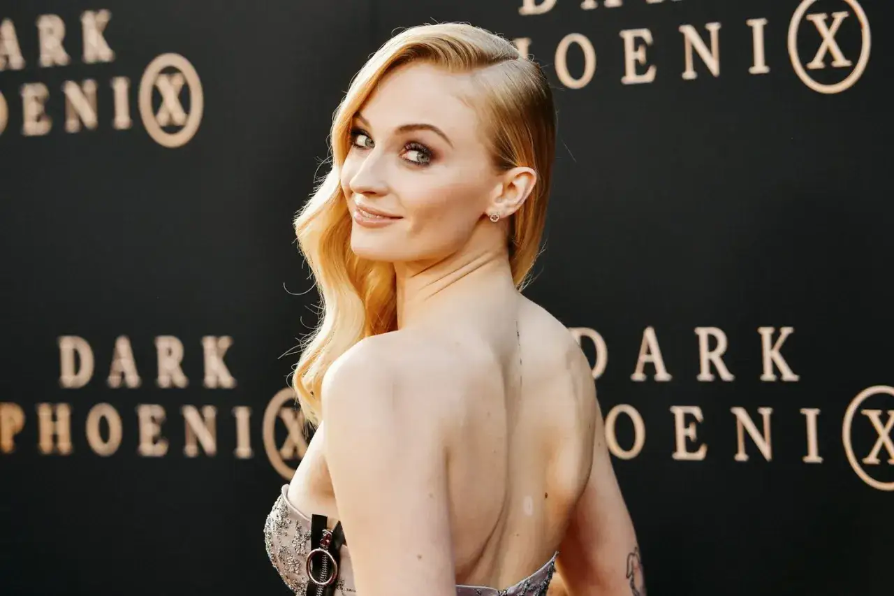 Sophie Turner, znana z filmów, seriali i programów, uśmiecha się na tle napisu
