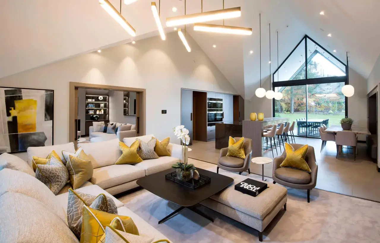 Sof&aacute; modular moderno con cojines dorados y grises. La sala de estar, ideal para como domotizar las luces de casa, tiene una chimenea, una cocina abierta y grandes ventanales.