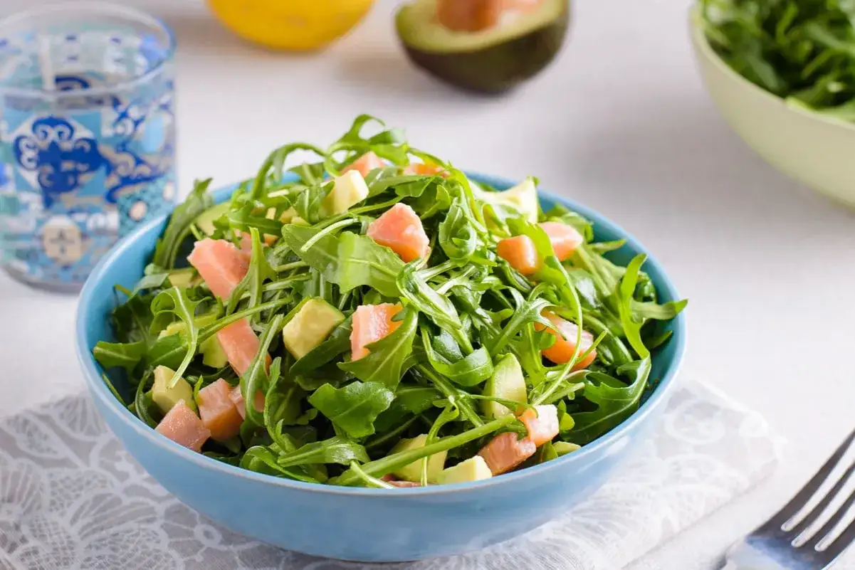 Insalata fresca con rucola, cubetti di avocado e pezzetti di salmone affumicato, servita in una ciotola blu.