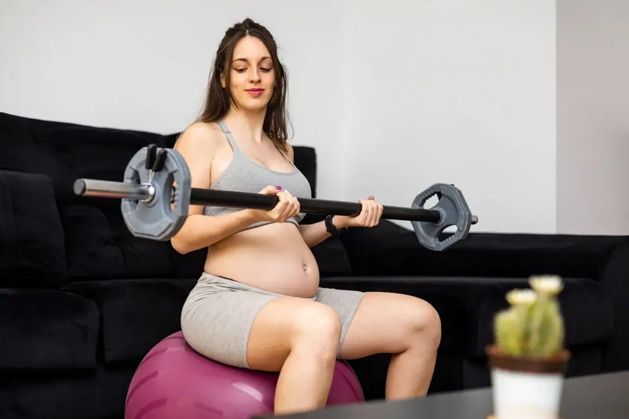 Gravid kvinna sitter p&aring; en tr&auml;ningsboll och lyfter en skivst&aring;ng. Hon &auml;r kl&auml;dd i tr&auml;ningskl&auml;der och har en tydlig gravidmage. Marklyft gravid &auml;r en &ouml;vning som kan anpassas.