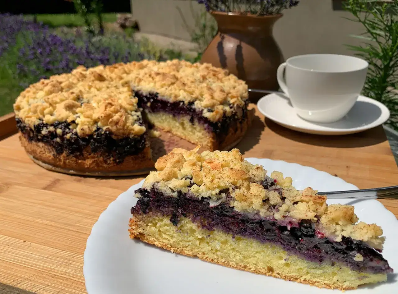 Ein saftiger Heidelbeerkuchen Landfrauenart mit Streuseln, daneben eine Tasse Kaffee und Lavendel im Hintergrund.