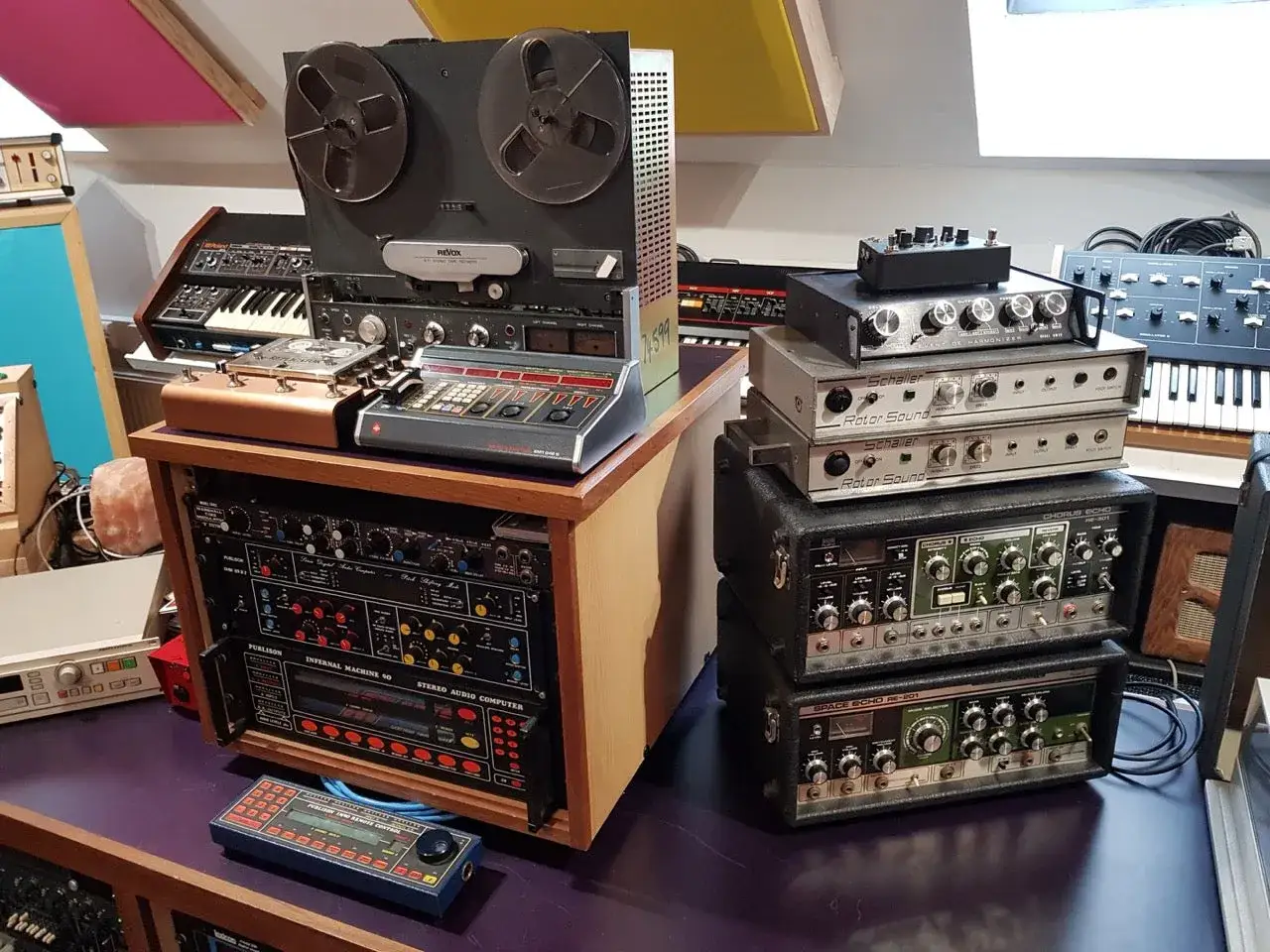A vintage studio setup with a Revox reel-to-reel tape machine, synthesizers, and rackmount effects units, including a little radiator-like device.