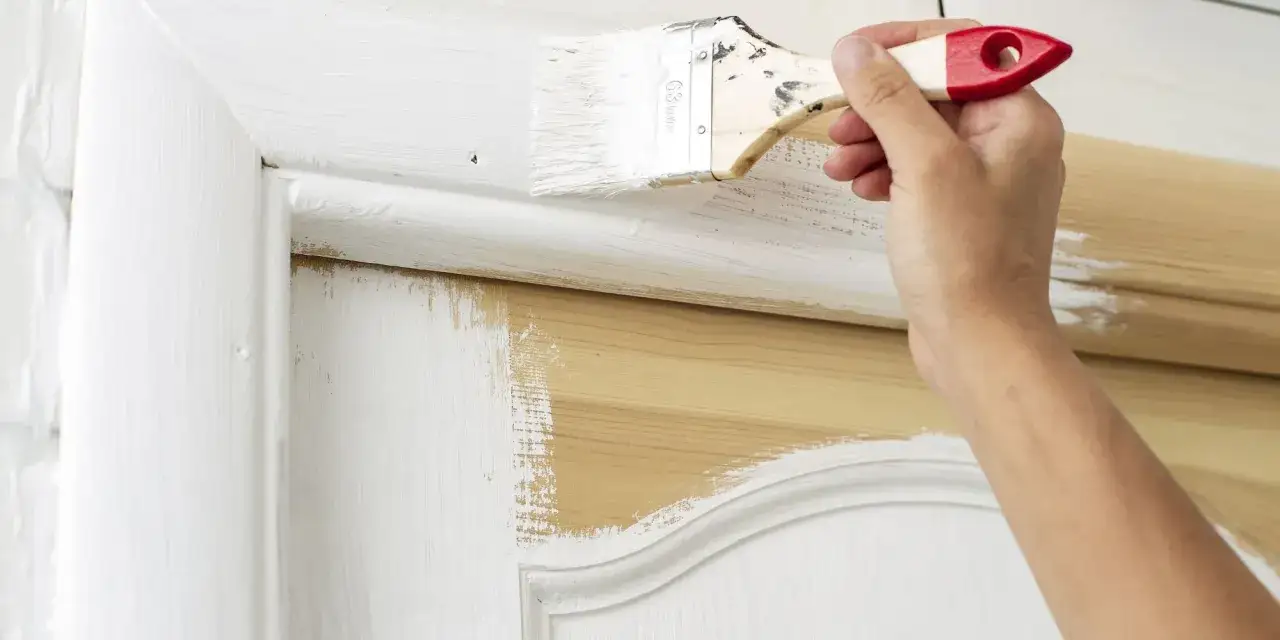 Main d'une personne peignant une porte en bois avec une peinture blanche, sans la dégonder. Le bois naturel apparaît par endroits.