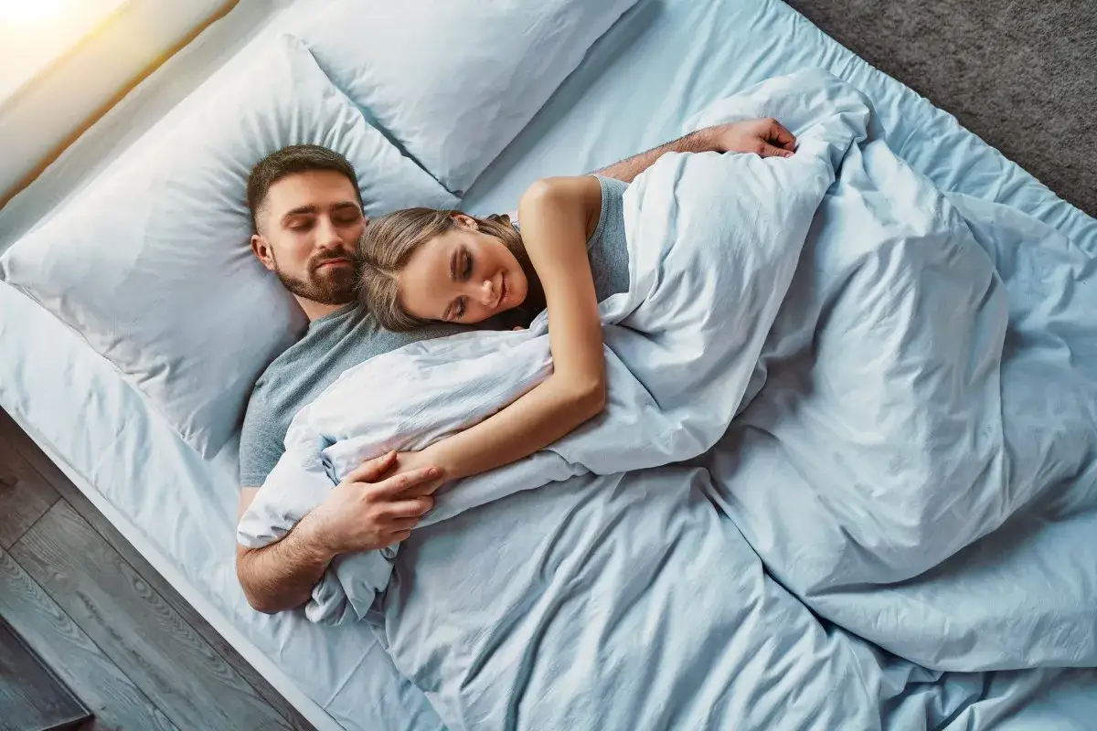 A couple sleeps peacefully, embracing under a white duvet. The woman rests her head on the man's chest, her hand on his arm. It's clear they sleep better next to someone they love.