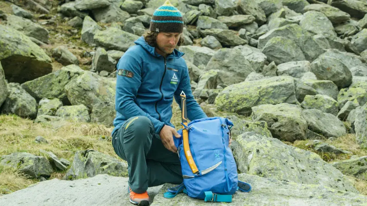 Bergsteiger prüft seine Packliste für die Hochtour. Ein blauer Rucksack und ein Eispickel liegen bereit.
