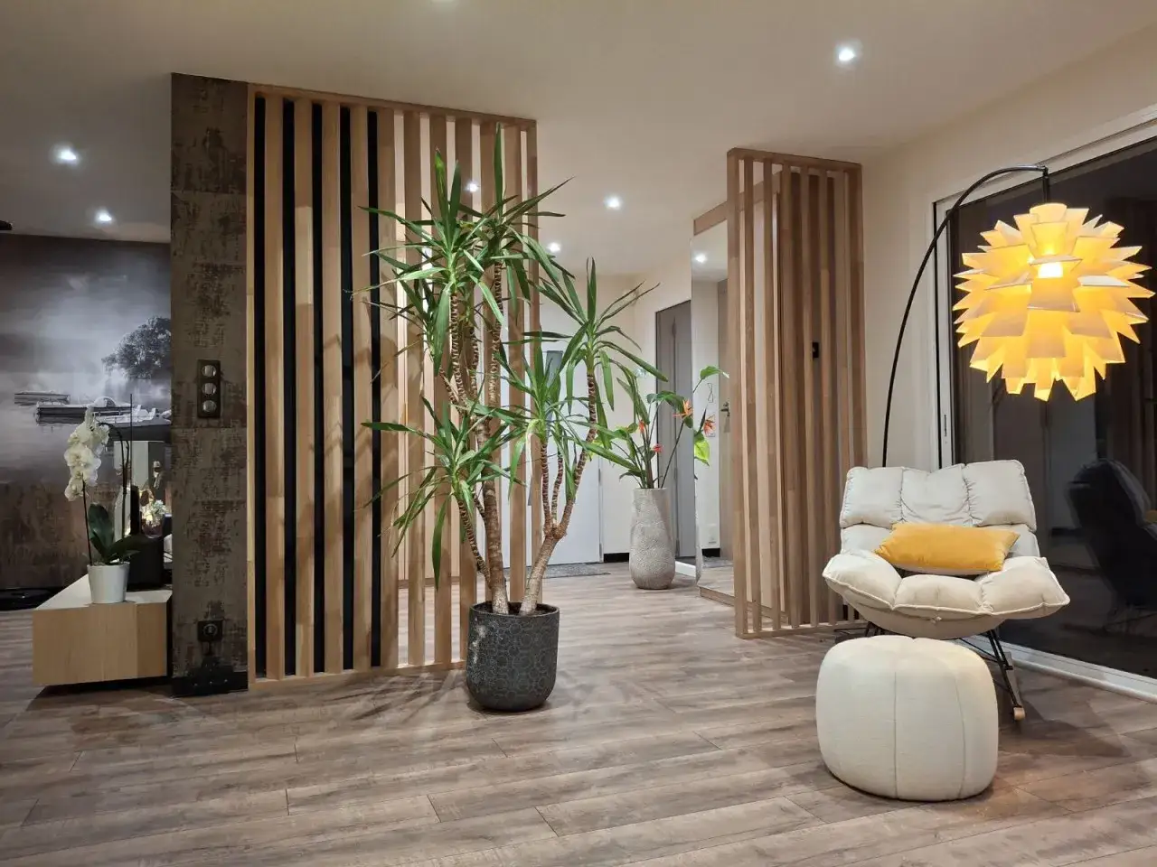 Un s&eacute;parateur en bois cr&eacute;e une entr&eacute;e dans le salon, avec une plante verte, un fauteuil confortable et une lampe design.