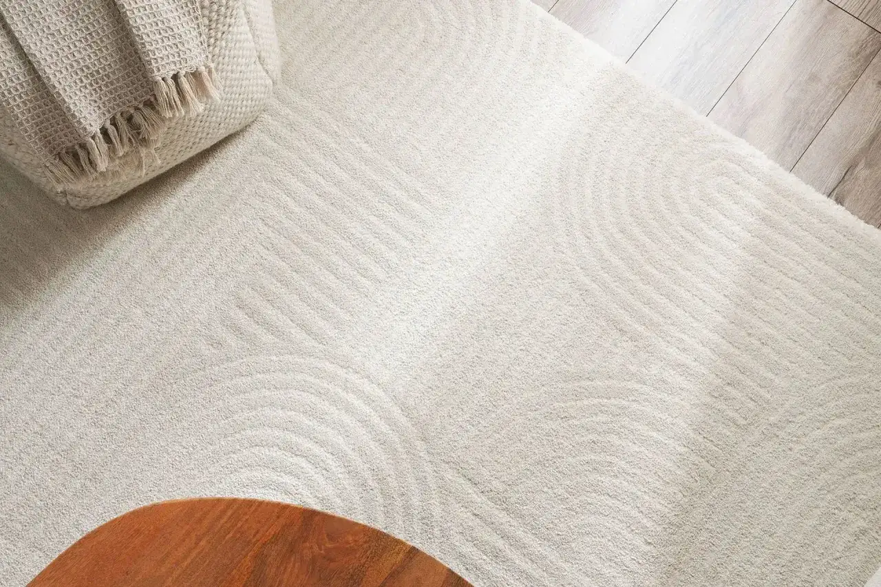 En ljus matta med vågformade mönster, en del av olika typer av mattor. En beige pläd med fransar ligger på en kudde i övre vänstra hörnet.