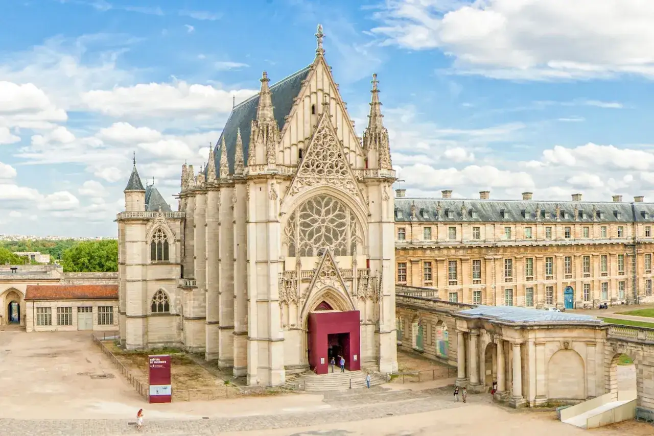 Majestuoso castillo de Vincennes con su imponente Sainte-Chapelle g&oacute;tica y edificios adyacentes bajo un cielo azul con nubes.