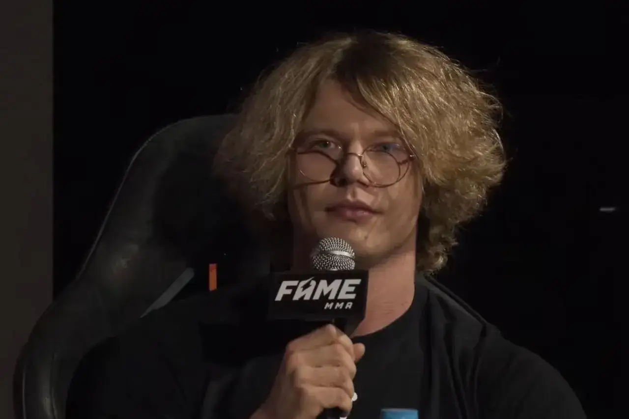 Natan Marcon, którego wiek jest tajemnicą, w okularach i z burzą blond włosów, trzyma mikrofon FAME MMA.