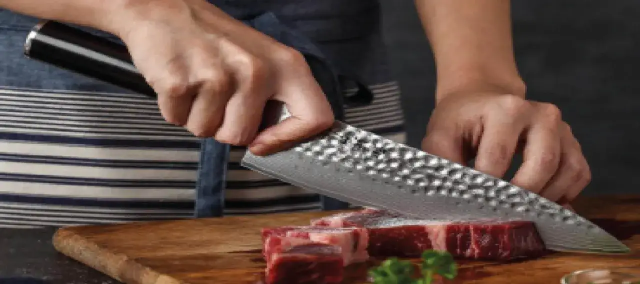 Manos cortando carne con un cuchillo con agujeros, de dise&ntilde;o martillado, sobre una tabla de madera.