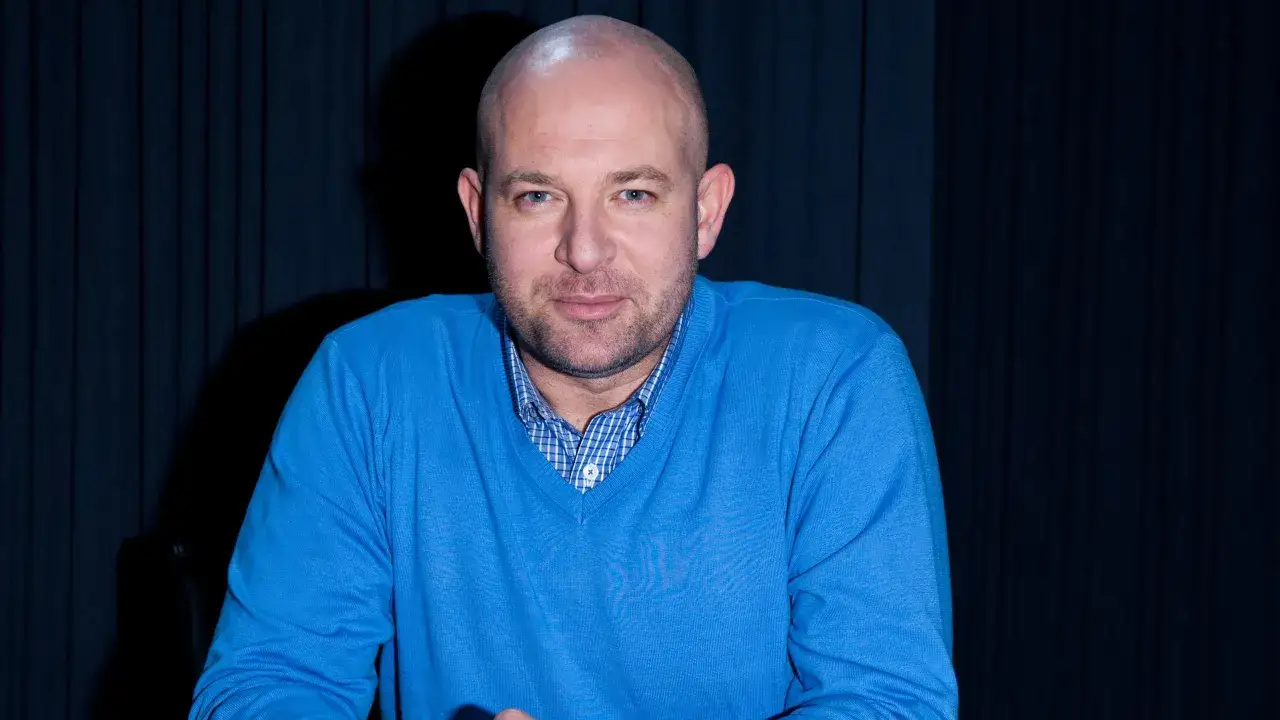 Szymon Bobrowski w niebieskim swetrze, znany z filmów, seriali i programów, pozuje na tle granatowej kurtyny.