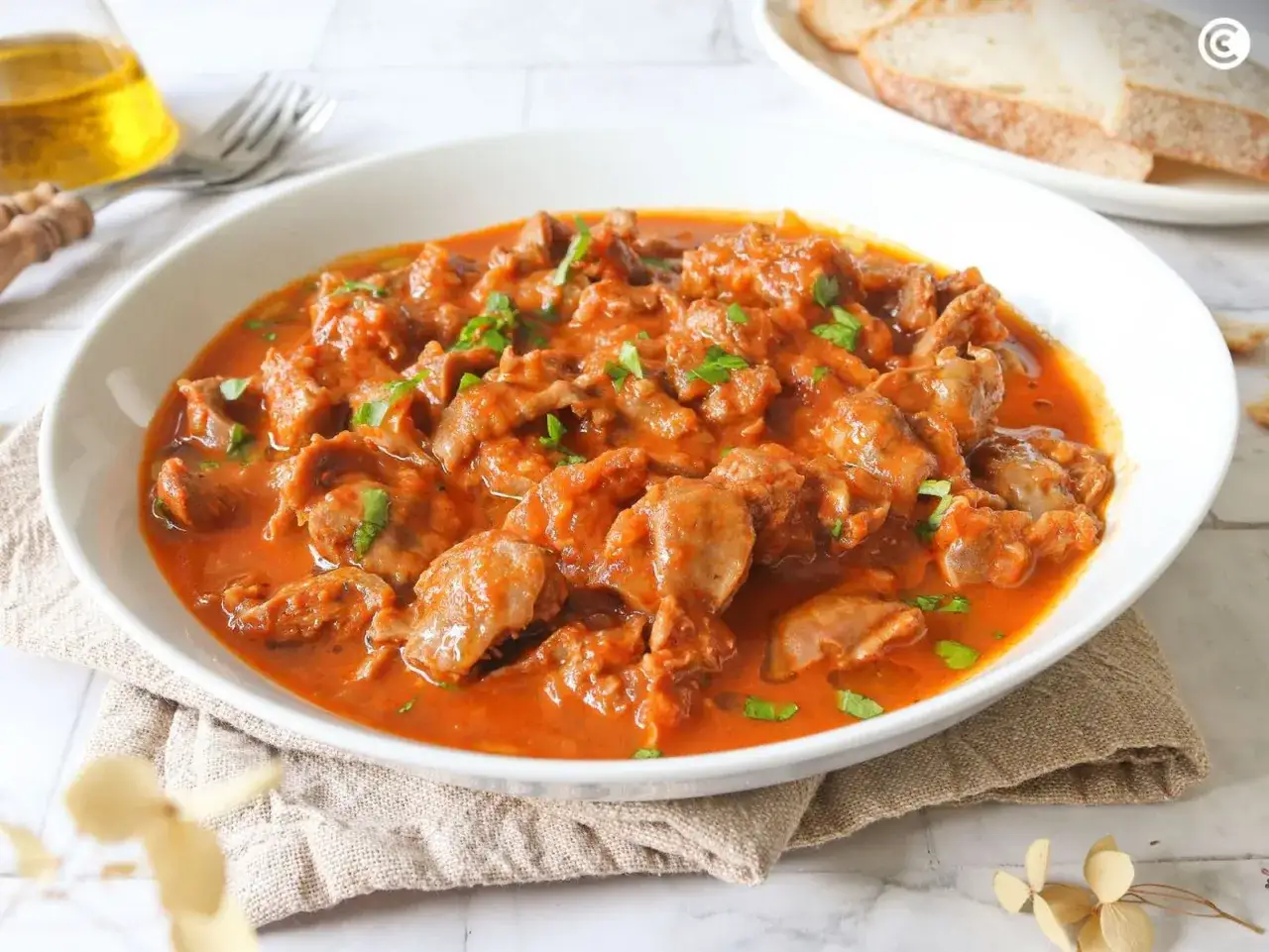 Plato de mollejas de pollo en salsa roja, decorado con perejil picado. Se sirve en un cuenco blanco sobre una servilleta de lino.