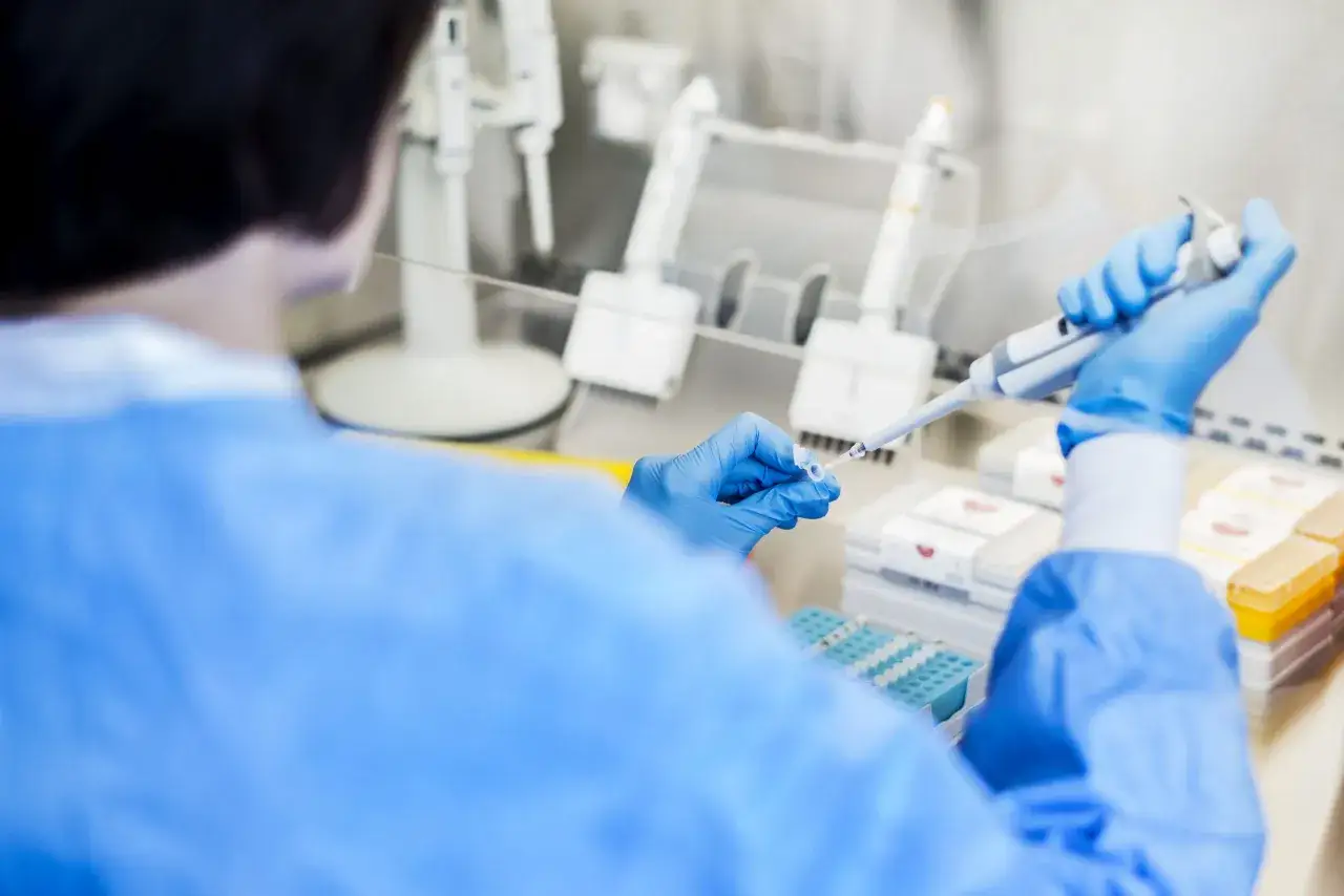 Naukowczyni w niebieskiej rękawiczce pobiera próbkę pipetą. W tle widać sprzęt laboratoryjny i pojemniki z odczynnikami. Allo MLR.