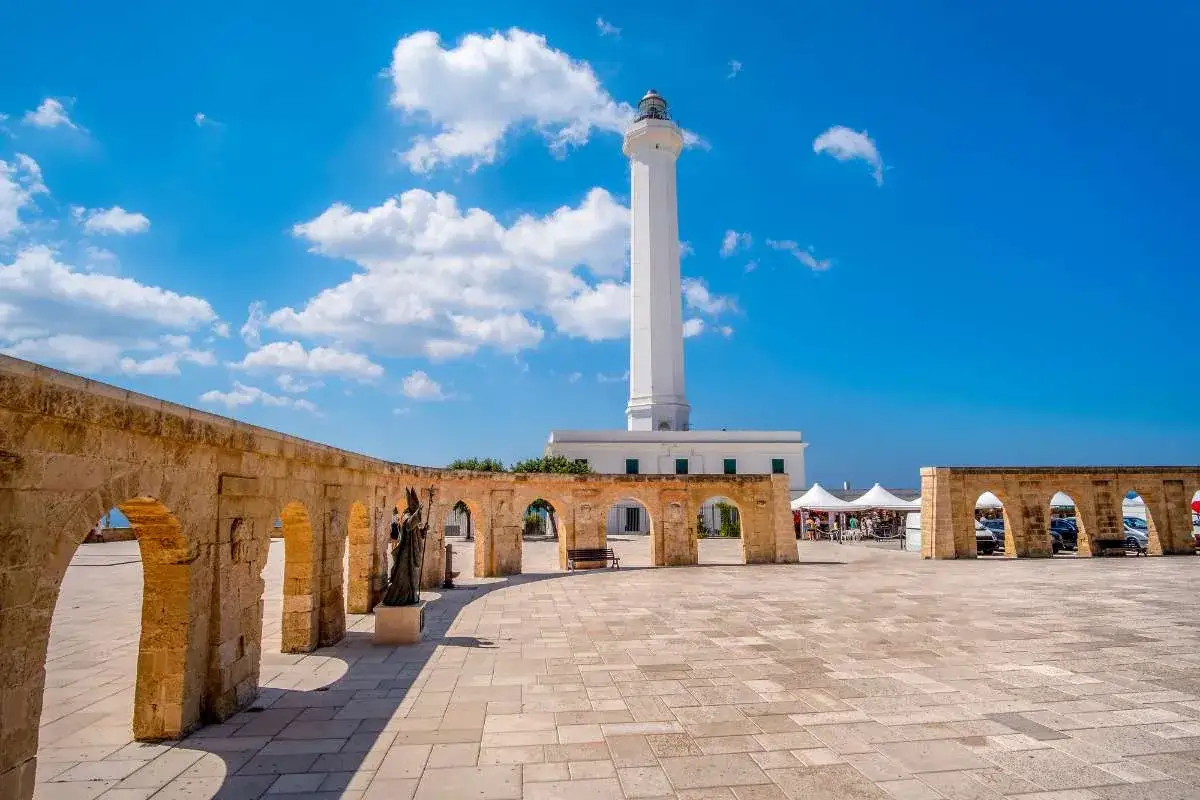 Santa Maria di Leuca: cosa vedere. Faro bianco imponente su piazza lastricata con arcate in pietra e una statua.