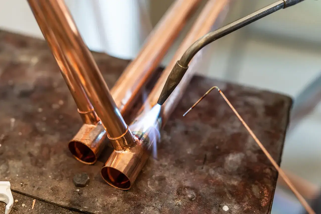 Un artisan réalise une soudure cuivre plomberie sur des tuyaux de cuivre, la flamme du chalumeau illuminant la scène.