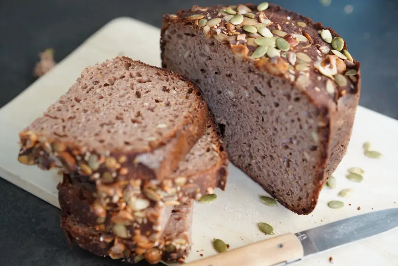 Pain de campagne à la farine de châtaigne, garni de graines de courge et de tournesol. Une tranche est coupée et empilée.