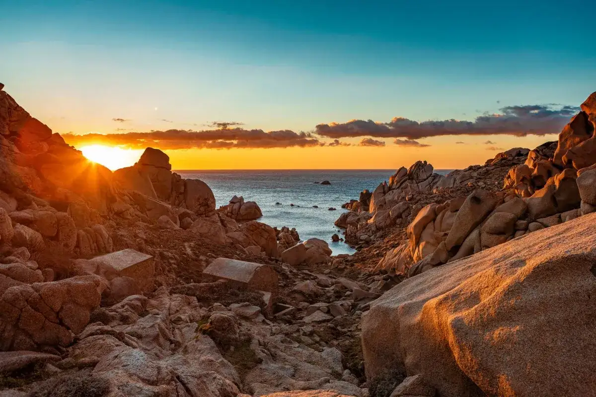 Sonnenuntergang am Capo Testa. Felsige Küste mit kleinen Steinhäusern, das Meer glitzert im warmen Licht.