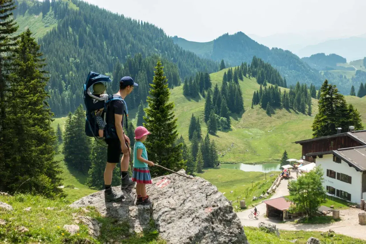 Vater mit Kind im Tragegestell und Tochter genie&szlig;en eine bergtour mit kindern. Herrliche Alpenlandschaft mit Bergsee und H&uuml;tte.
