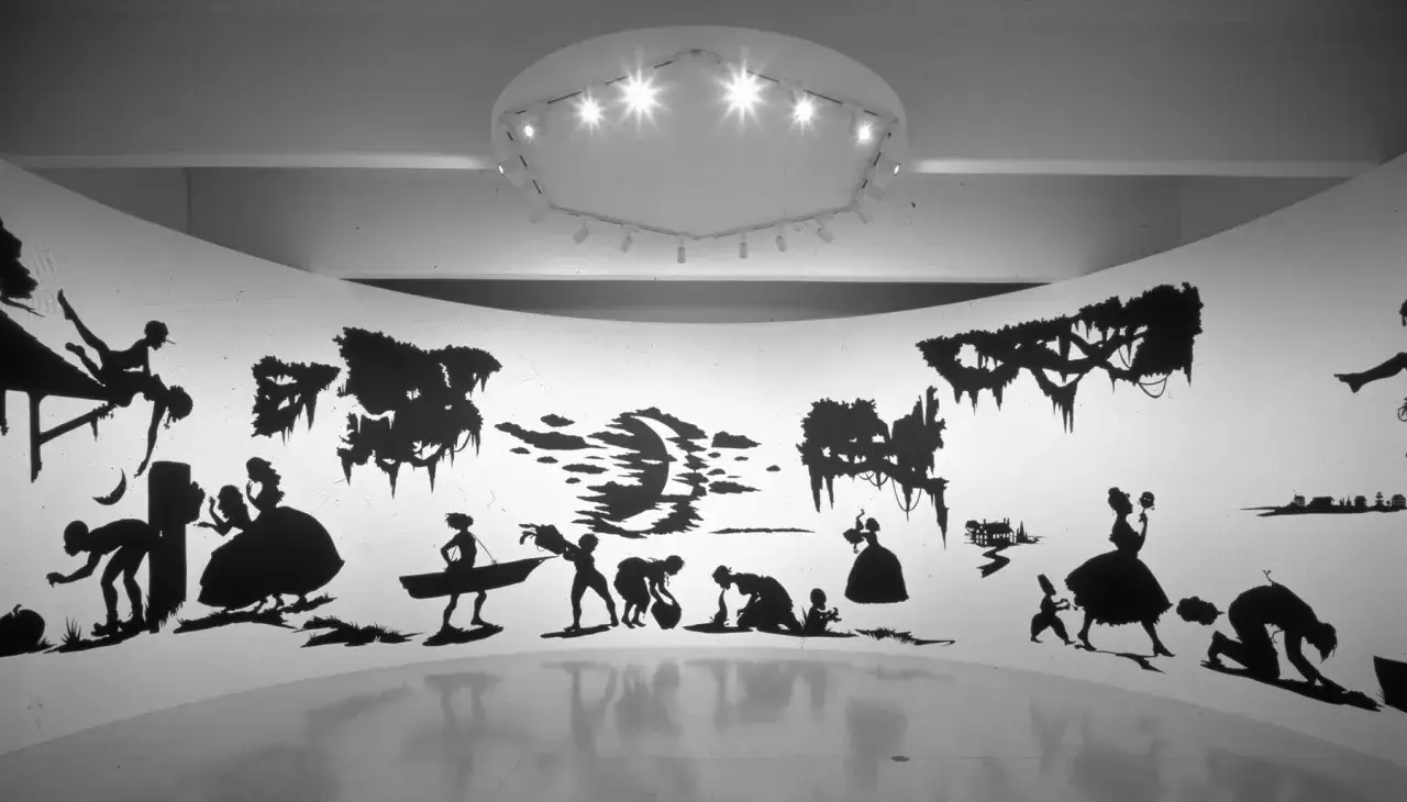 A panoramic Kara Walker artwork installation features black silhouettes of figures in historical dress, some engaged in labor, others in leisure, set against a stark white background with shadowy trees.