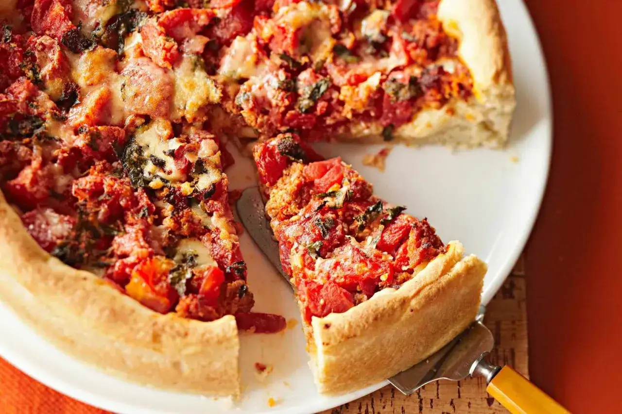En saftig deep dish pizza med tjock botten, fylld med tomater, ost och örter. En bit lyfts upp med en spade.