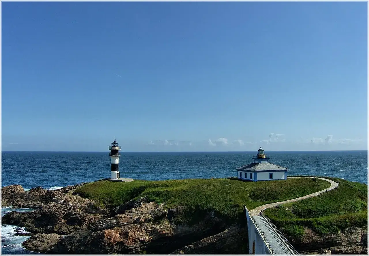 El faro de Ribadeo, con su torre a rayas y una casa blanca, vigila el mar azul bajo un cielo despejado.