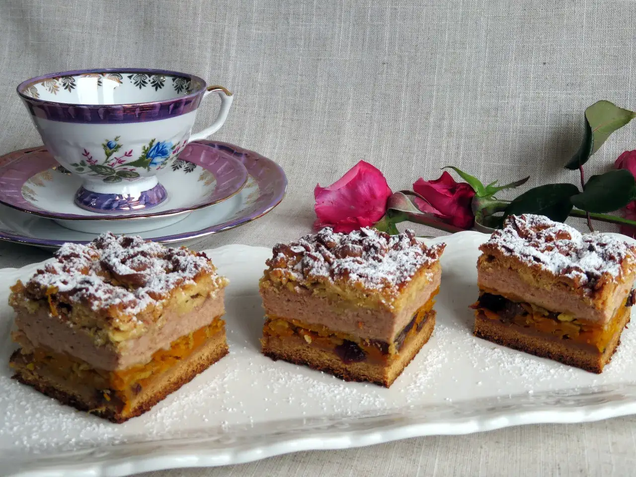 Trzy kawałki pysznego ciasta na jesień, posypane cukrem pudrem, obok eleganckiej filiżanki i róż.