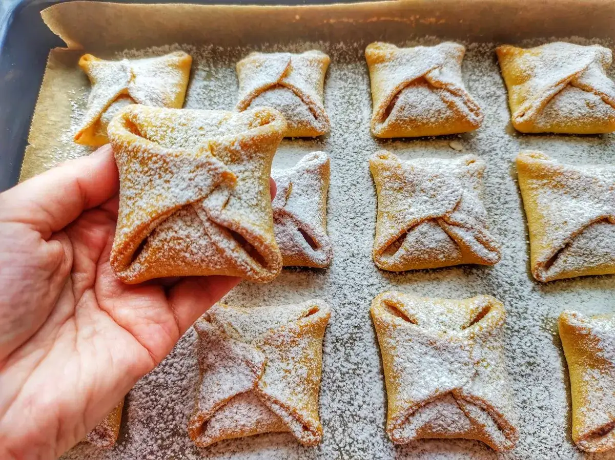 Ręka trzyma jedno z wielu pysznych, kwadratowych ciastek z jabłkami, posypanych cukrem pudrem.