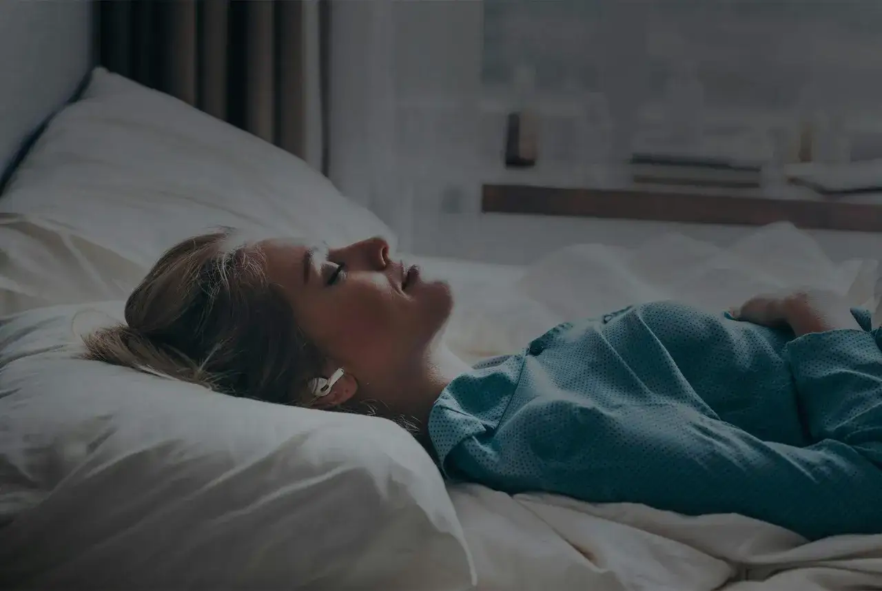 En kvinna ligger i sängen med hörlurar, ansiktet vänt uppåt, kanske upplever ångest på morgonen.