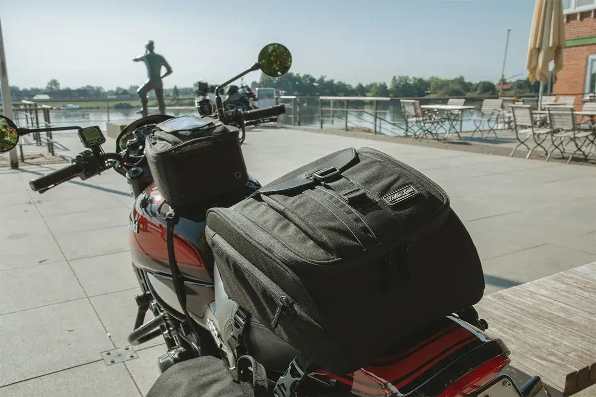Equipaje en moto listo para la aventura. Una bolsa negra sobre el asiento, con un paisaje de río y mesas al fondo.