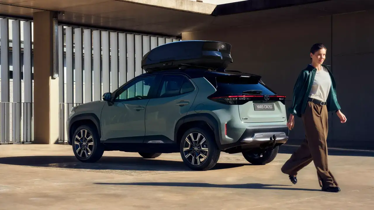 Ein stilvoller Yaris Cross 2026 mit Dachbox, bereit f&uuml;r Abenteuer. Eine Frau geht l&auml;ssig daran vorbei.