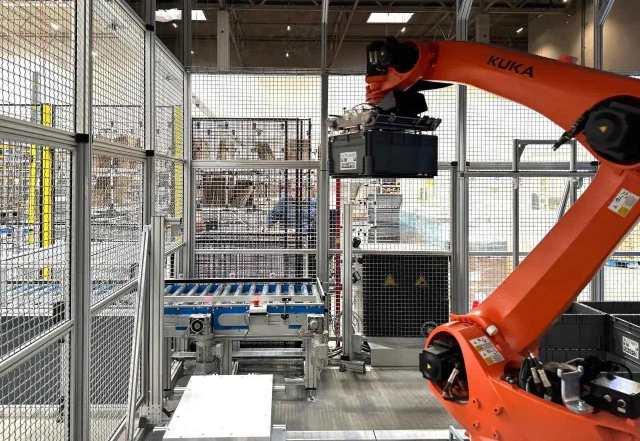 Pomarańczowy robot KUKA podnosi czarną skrzynkę. To jak serial, gdzie każdy odcinek to nowy etap produkcji.