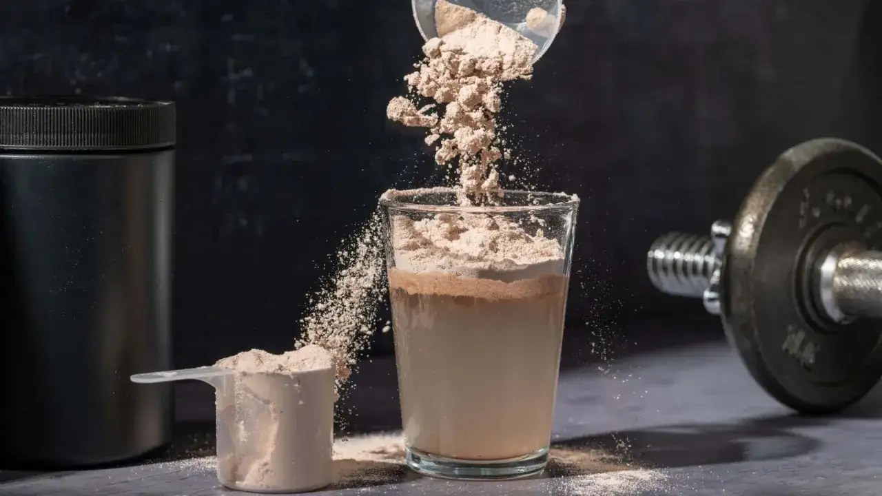 Pulver rieselt in ein Glas mit Flüssigkeit, um einen proteinshake mit milch oder wasser zuzubereiten. Daneben ein Löffel mit Pulver und eine Hantel.