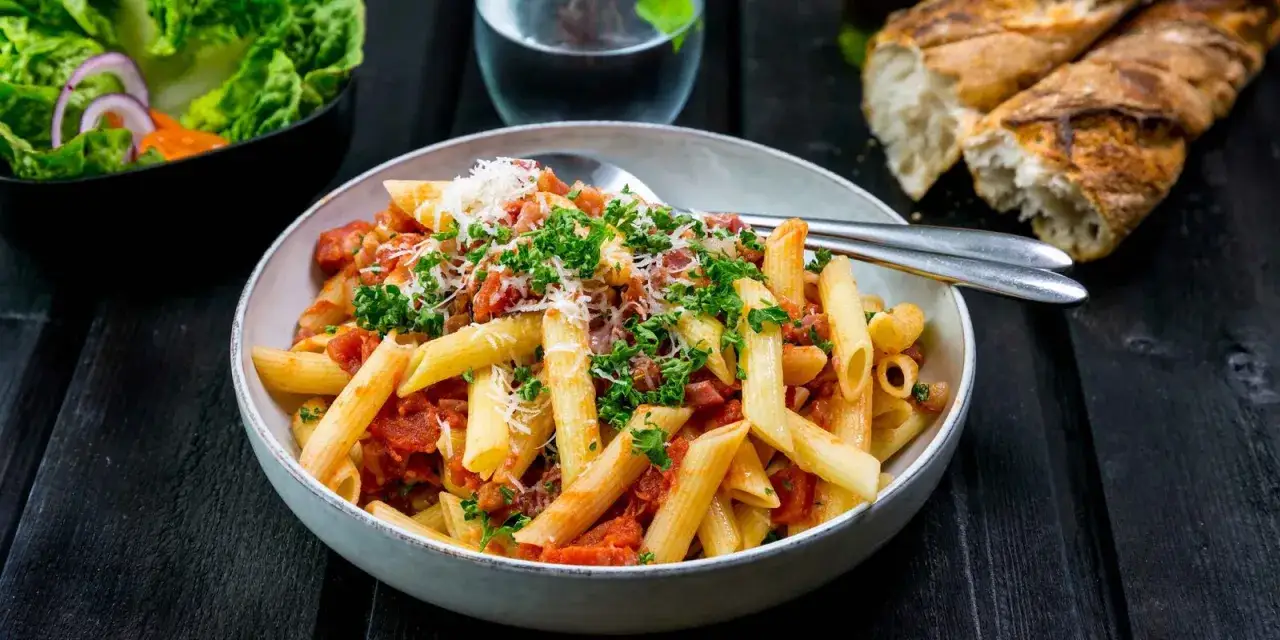 En sk&aring;l med penne all'arrabiata, toppad med persilja och parmesan. Bredvid en sallad och ett glas vatten.
