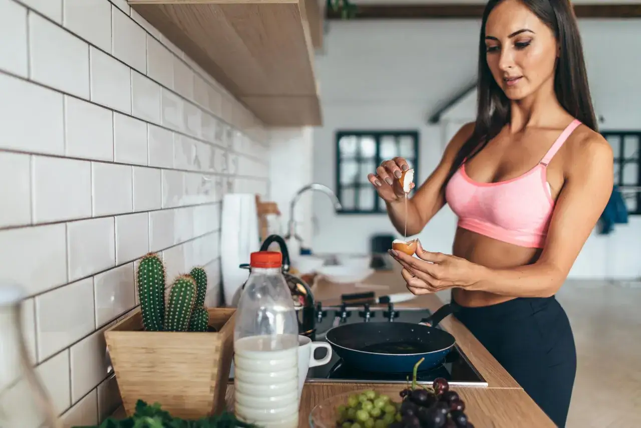 Kobieta w sportowym topie przygotowuje jajka na patelni, planując cardio na czczo. Na blacie stoją kaktusy i owoce.