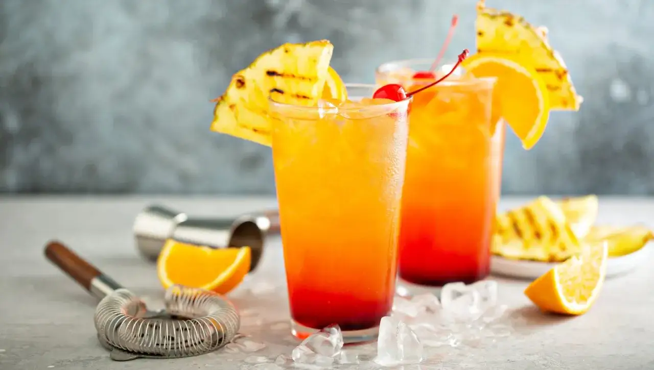 Dwa drinki tequila sunrise z lodem, plasterkami pomarańczy i ananasa, ozdobione wiśnią. Idealne na letnie wieczory.