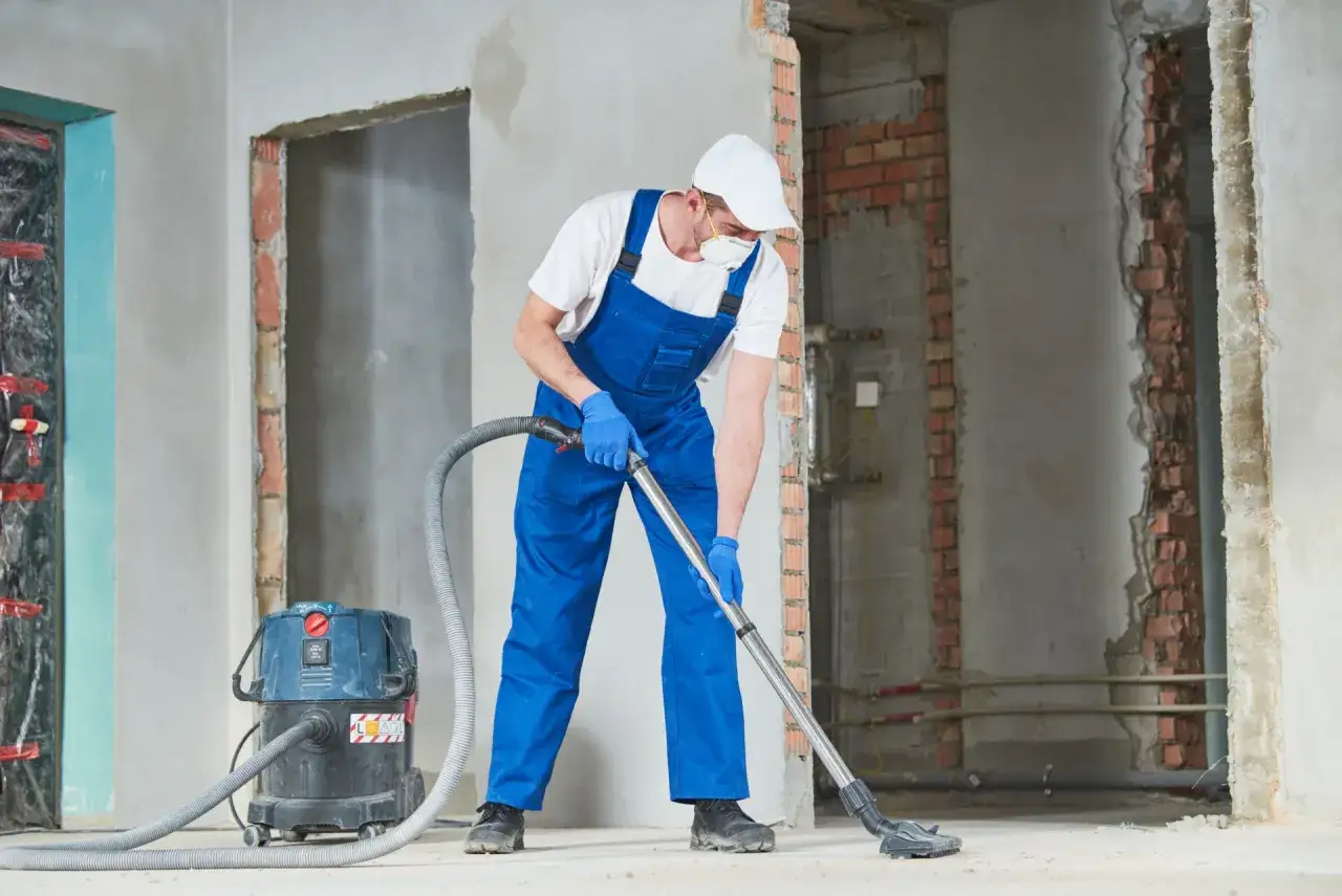 Un ouvrier en combinaison bleue utilise un aspirateur pour nettoyer le mur après ponçage dans un chantier.