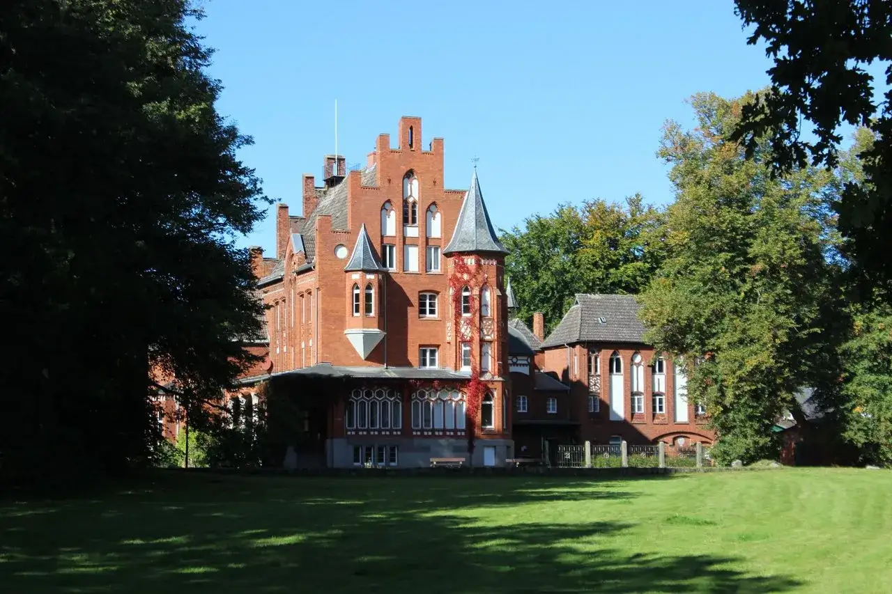 Das Schloss Kalkhorst, ein roter Backsteinbau mit Türmchen und Erkern, steht auf einer grünen Wiese, umgeben von Bäumen.