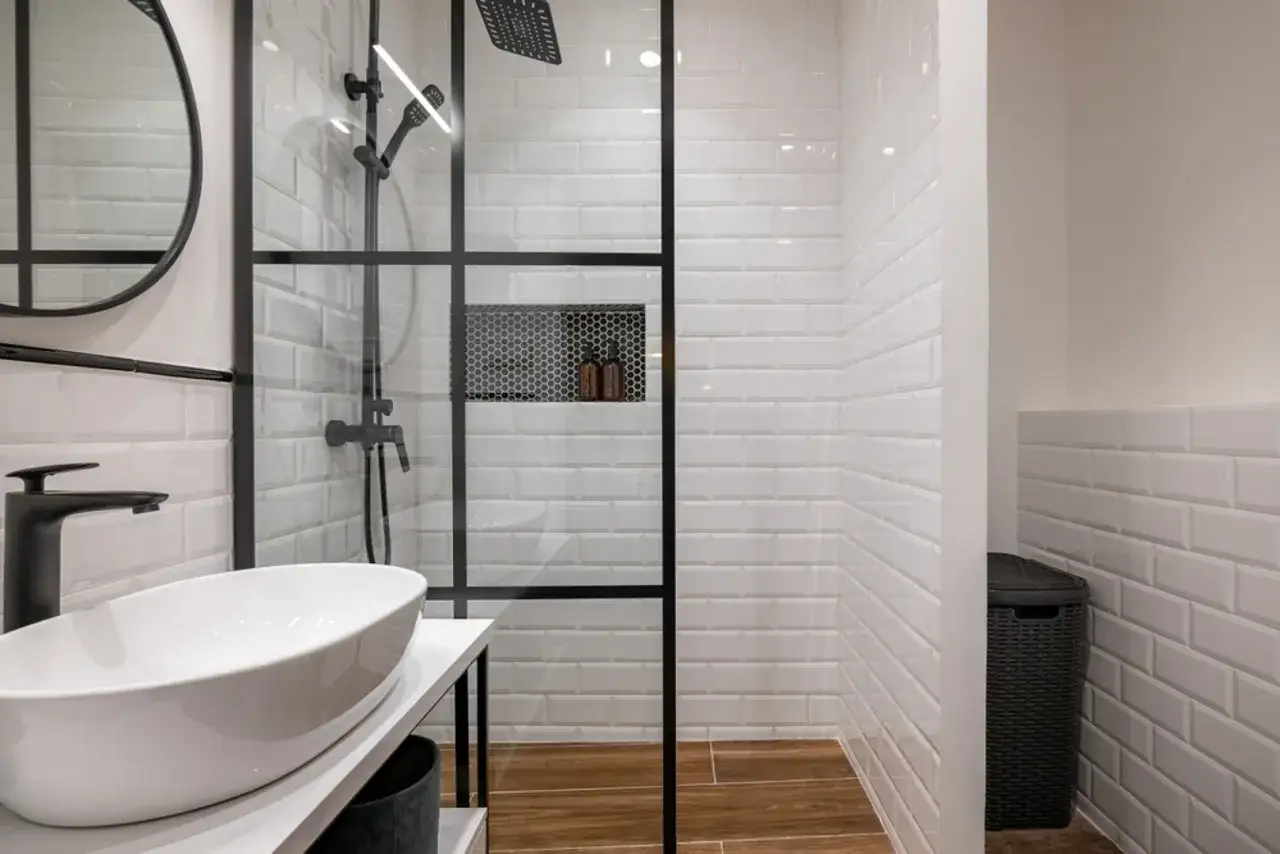 Modernos ba&ntilde;os con pared en la ducha de azulejos blancos y suelo de madera. Lavabo sobre mueble y grifer&iacute;a negra.