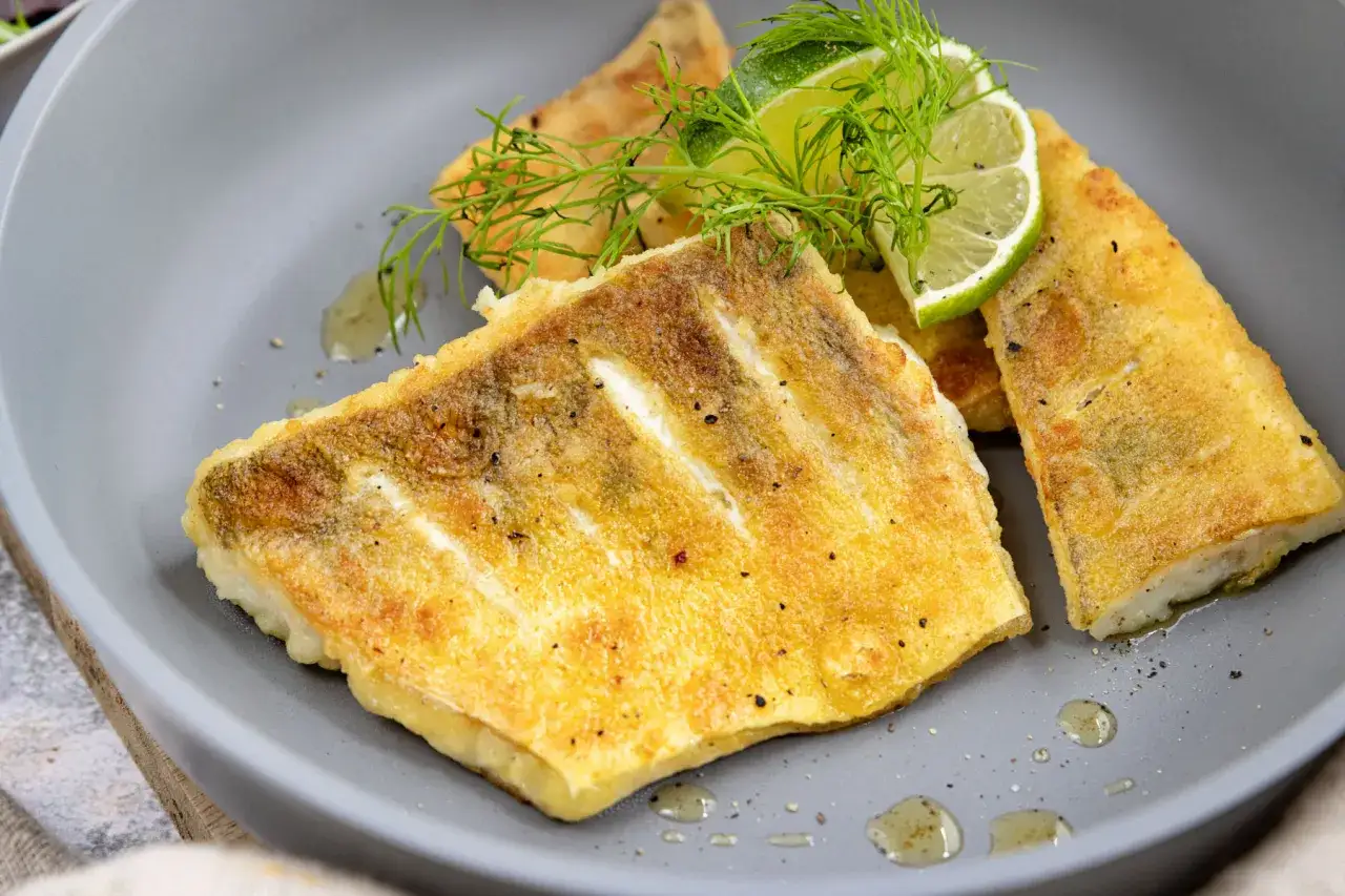 Goldbraun gebratenes Zanderfilet ohne Haut, garniert mit Dill und Limettenspalten, auf einem grauen Teller.