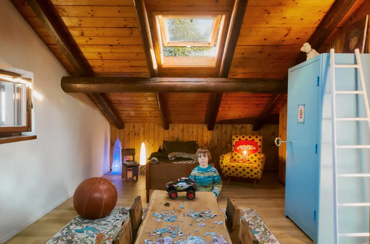 Bambino in maglione colorato gioca con un'auto giocattolo su un tavolo con puzzle. La stanza ha un tetto spiovente in legno, un armadio azzurro e una poltrona gialla a pois. Un'atmosfera da 