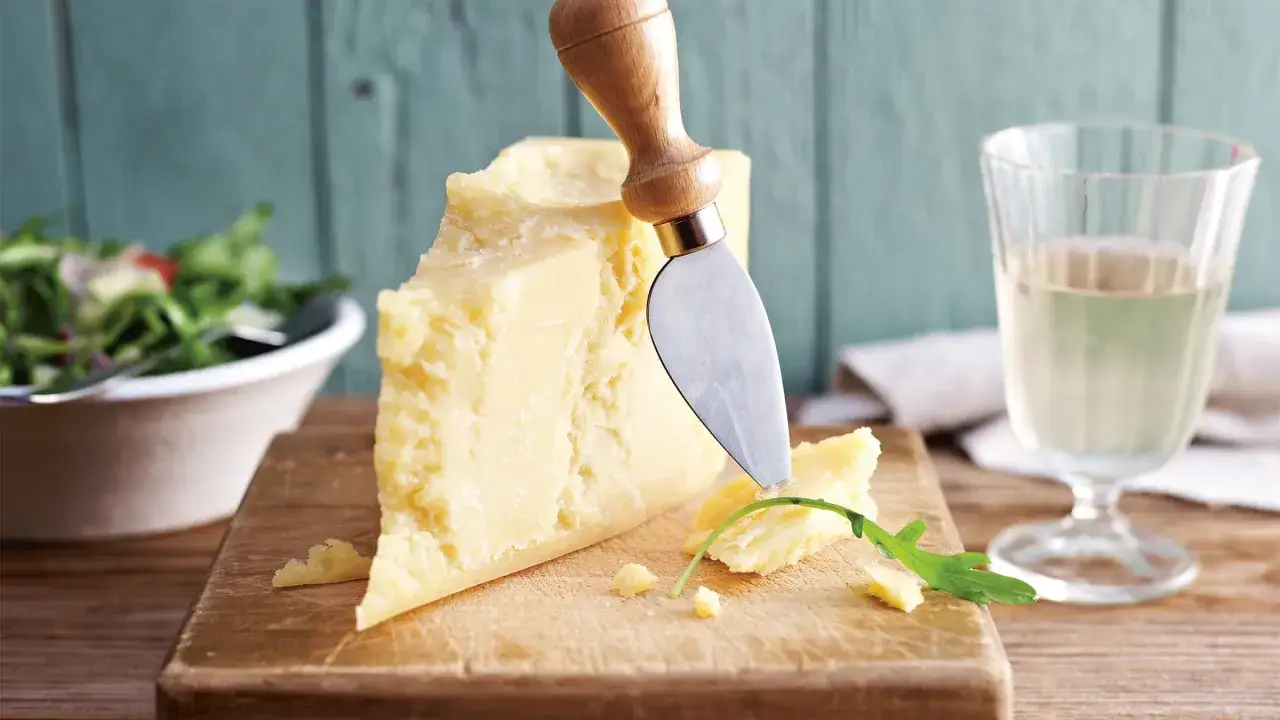 Ein Stück Parmesan, vegetarisch, wird mit einem Messer auf einem Holzbrett angeschnitten. Daneben ein Salat und ein Glas Wein.