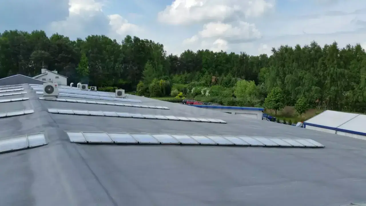 Szary, lekki dach płaski z rzędami świetlików i klimatyzatorami, otoczony zielenią drzew.